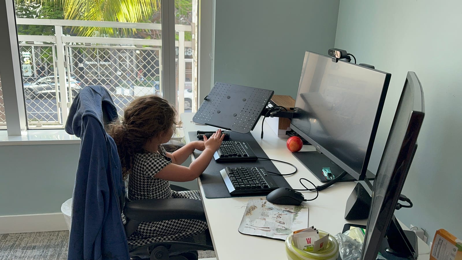 A toddler sitting in the desk chair of a home office, reaching up to type on the keyboard between two monitors and a laptop stand.